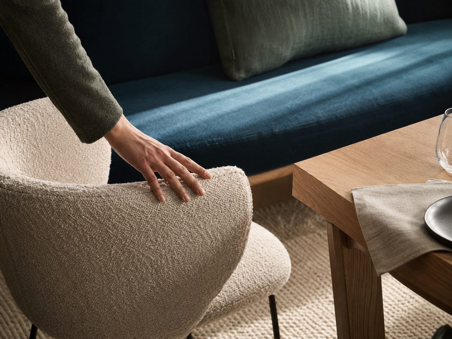Main posée sur une assise en tissu bouclé écru, banquette en velours bleu et table en bois clair en arrière-plan.